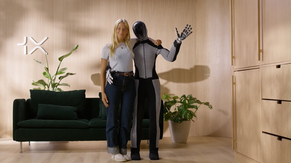 1X's EVE wheeled humanoid robot standing next to a person in an office setting