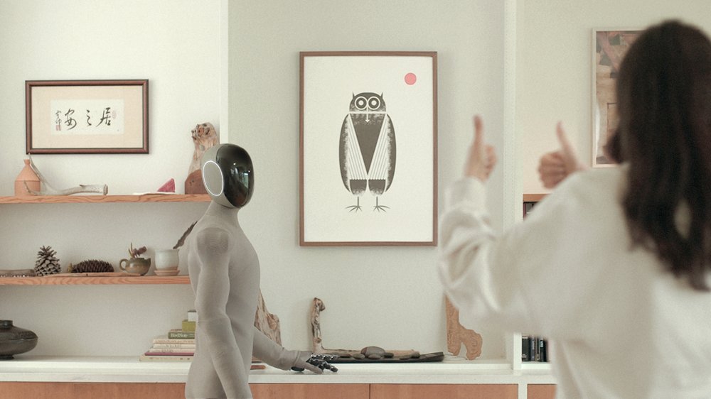 1X's NEO humanoid robot standing in a home living room, giving a thumbs-up gesture to a person